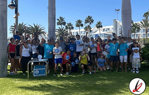 Grupo de personas, adultos y niños, participando en un evento al aire libre en Lanzarote, con palmeras y edificios modernos de fondo.