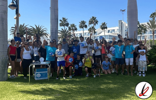 Grupo de personas, adultos y niños, participando en un evento al aire libre en Lanzarote, con palmeras y edificios modernos de fondo.