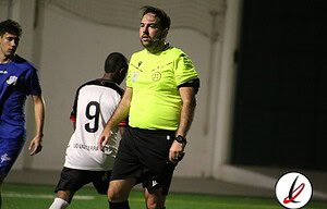 Árbitro de fútbol con camiseta neón, en un estadio, con jugadores en el fondo durante un partido.