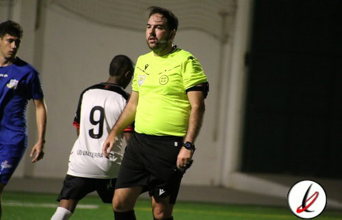 Árbitro de fútbol con camiseta neón, en un estadio, con jugadores en el fondo durante un partido.