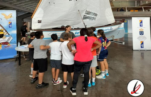 Barco de vela en exhibición en una feria deportiva con niños y una monzotriz que explica detalles, en un espacio interior con otros stands y logotipos en el fondo.