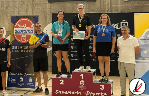 1. Jugadores en el podio de un torneo de tenis de mesa en Lanzarote, con medallas y trofeos, fondos con logos de patrocinadores y público en el gimnasio.