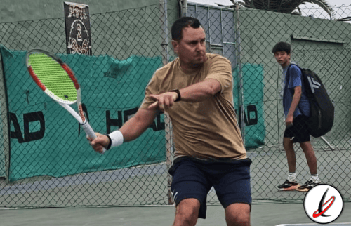 Jugador de tenis en acción en una cancha con malla, sosteniendo una raqueta y preparándose para golpear la pelota durante un partido en Lanzarote Deportivo.