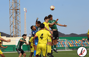 Un grupo de futbolistas juveniles saltando para cabecear el balón en un partido de fútbol en un campo de césped artificial, con gradas y cielo despejado de fondo.
