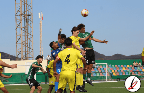 Un grupo de futbolistas juveniles saltando para cabecear el balón en un partido de fútbol en un campo de césped artificial, con gradas y cielo despejado de fondo.
