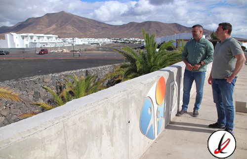 Jugadores conversando junto a un mural en Lanzarote con fondo de volcanes y ciudad, en un entorno al aire libre con cielo despejado y clima soleado, en un día caluroso.