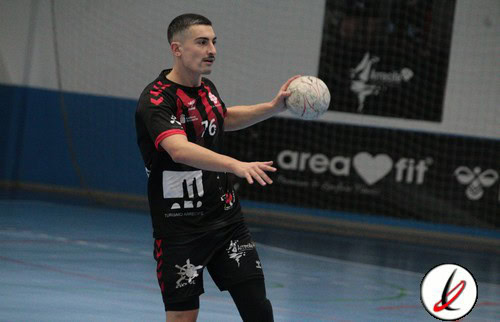 Un jugador de fútbol sala en acción, preparado para pasar el balón en una pista cubierta de deporte, con uniforme negro y detalles rojos, dentro de un estadio deportivo.