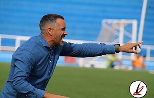 Entrenador en la banda de un campo de fútbol, dando instrucciones durante un partido en un estadio con gradas azules.
