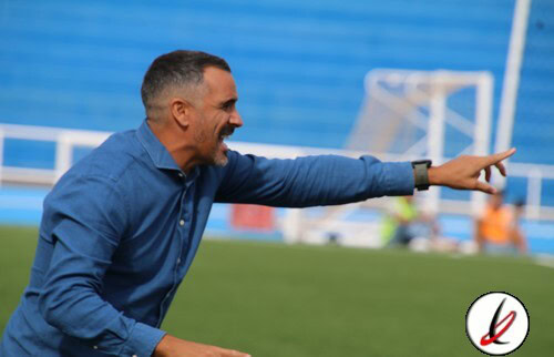 Entrenador en la banda de un campo de fútbol, dando instrucciones durante un partido en un estadio con gradas azules.