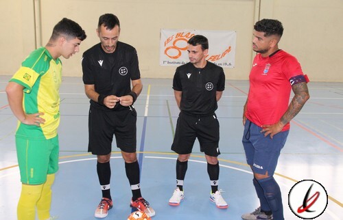 1. Dos jugadores de fútbol, uno en uniforme amarillo y verde y otro en uniforme azul marino, junto a dos árbitros en camiseta negra, en una cancha cubierta de fútbol sala.