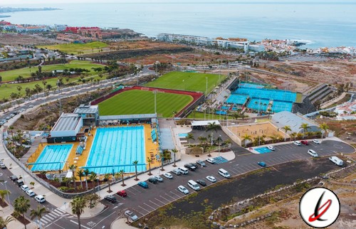 Bañera de piscina al aire libre en Lanzarote, con espectadores y zonas de descanso, vista aérea de instalaciones deportivas en un entorno costero con playa y mar al fondo.