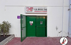 Puerta de entrada a la sede del C.B. Mace Tías, con cartel del club y puerta metálica verde, en una pared blanca, en Lanzarote, ideal para promover el deporte y el baloncesto en la isla.