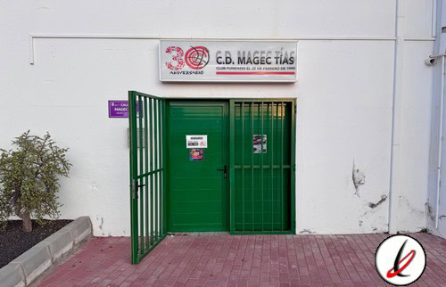 Puerta de entrada a la sede del C.B. Mace Tías, con cartel del club y puerta metálica verde, en una pared blanca, en Lanzarote, ideal para promover el deporte y el baloncesto en la isla.