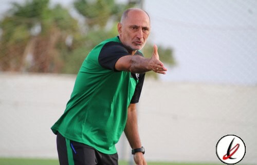 Entrenador dando instrucciones en un campo de fútbol, con fondo de muro y árboles, durante un entrenamiento o partido.