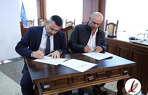 1. Dos hombres en una reunión formal firmando documentos en una mesa de madera, con bandera de la Unión Europea y bandera de España visibles en el fondo, en una oficina con decoración moderna.