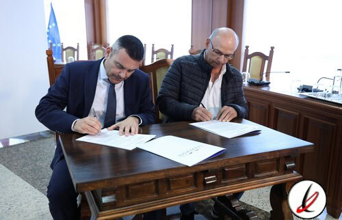 1. Dos hombres en una reunión formal firmando documentos en una mesa de madera, con bandera de la Unión Europea y bandera de España visibles en el fondo, en una oficina con decoración moderna.