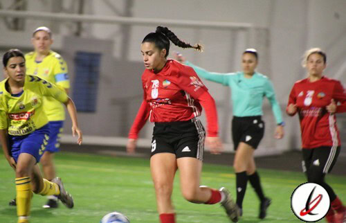 1. Jugadora de fútbol femenino en acción durante un partido en el campo, con otras jugadoras y árbitras en el fondo.