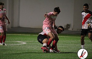 Jugador de fútbol en acción en el estadio Lanzarote Deportivo, durante un partido. El futbolista lleva un uniforme rosa y está evitando que el oponente en camiseta blanca y negra controle el balón en el campo artificial.