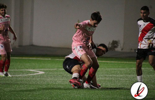Jugador de fútbol en acción en el estadio Lanzarote Deportivo, durante un partido. El futbolista lleva un uniforme rosa y está evitando que el oponente en camiseta blanca y negra controle el balón en el campo artificial.