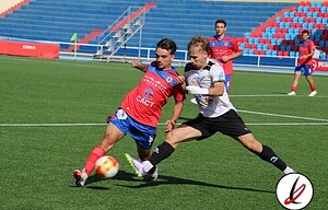Lucha por el balón entre dos futbolistas en un partido en un estadio de Lanzarote, con público en las gradas y césped bien cuidado. Jugadores en acción durante un partido de fútbol en un campo profesional.