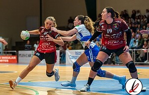 1. Tres jugadoras de balonmano luchan por el balón en una pista cubierta durante un partido femenino. Jugadoras en uniformes deportivos rojos y azules en acción intensa con público al fondo.