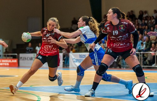 1. Tres jugadoras de balonmano luchan por el balón en una pista cubierta durante un partido femenino. Jugadoras en uniformes deportivos rojos y azules en acción intensa con público al fondo.