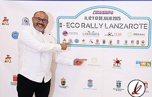 1. Persona en una reunión oficial sosteniendo un cartel del Eco Rally Lanzarote 2025, con logotipos y patrocinadores en el fondo, vestido con camisa blanca y gafas, en un ambiente formal y festivo.