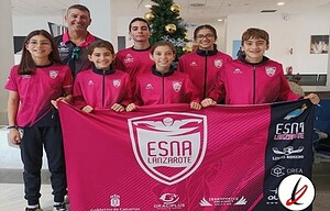 Jugadoras de fútbol infantil con su entrenador, sosteniendo una bandera del equipo ESNA Lanzarote, en un interior bien iluminado, con rostros sonrientes y vestimenta deportiva rosa con el escudo del equipo.