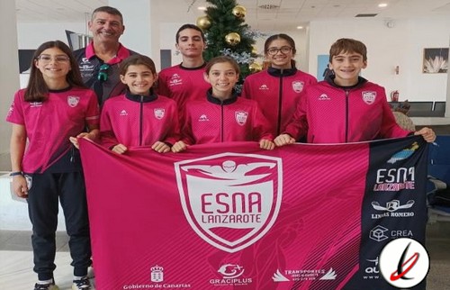 Jugadoras de fútbol infantil con su entrenador, sosteniendo una bandera del equipo ESNA Lanzarote, en un interior bien iluminado, con rostros sonrientes y vestimenta deportiva rosa con el escudo del equipo.