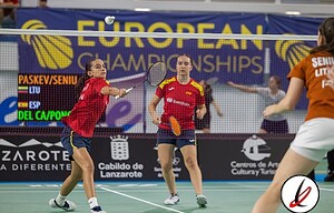 1. Jugadoras de bádminton en acción durante un partido en Lanzarote, con fondo de cartel de UEFA European Championships y público en las gradas.