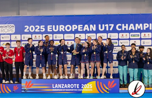 La imagen muestra a un grupo de jugadores de bádminton juvenil celebrando en el podio del Campeonato Europeo Sub-17 2025 en Lanzarote, con uniforme azul y varios jugadores sosteniendo medallas, en un escenario con cartel de fondo y logotipos de patrocinadores.