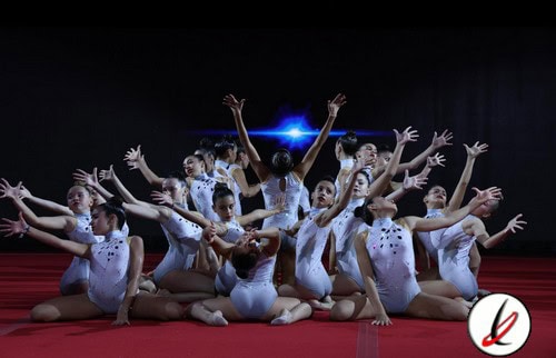 1. Grupo de gimnastas en danza artística en escenario con luces azules y fondo negro, todos con trajes blancos y posiciones coordinadas.