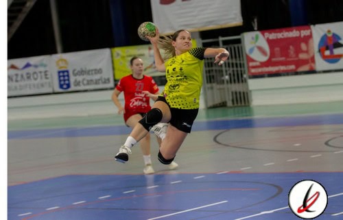Una jugadora de balonmano en acción en un partido, con otra jugadora en el fondo en un pabellón deportivo, vistiendo uniforme amarillo y rojo, con público y anuncios en la pista.