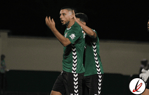 Un futbolista celebrando un gol en un partido nocturno, vestido con camiseta verde y pantalones cortos, en un estadio.