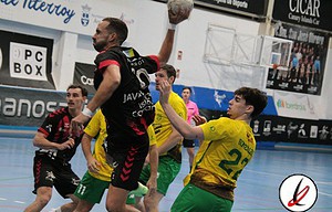 Tiro enuncidad de balonmano en un partido, jugador con camiseta negra saltando con balón en mano, defensores con camisetas amarillas y verdes en pista cubierta, en interior de pabellón deportivo.