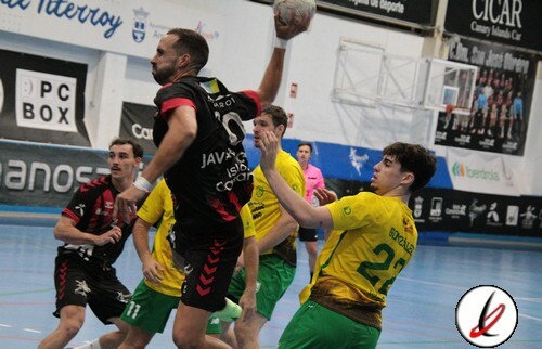 Tiro enuncidad de balonmano en un partido, jugador con camiseta negra saltando con balón en mano, defensores con camisetas amarillas y verdes en pista cubierta, en interior de pabellón deportivo.