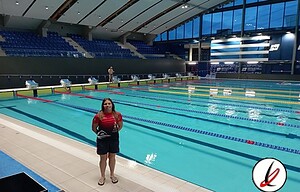 Natación en una piscina cubierta con equipada para entrenamiento y competición, con azulejos azules y grada para espectadores en el fondo, y una nadadora en primer plano con uniforme rojo y pantalones cortos.