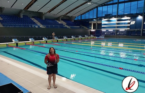 Natación en una piscina cubierta con equipada para entrenamiento y competición, con azulejos azules y grada para espectadores en el fondo, y una nadadora en primer plano con uniforme rojo y pantalones cortos.