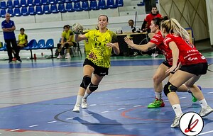 Lanzadora de balonmano en acción durante un partido en un pabellón deportivo, con jugadoras en movimiento y público observando en las gradas.
