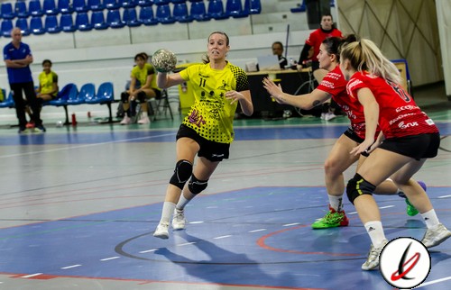 Lanzadora de balonmano en acción durante un partido en un pabellón deportivo, con jugadoras en movimiento y público observando en las gradas.