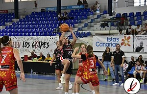 Una jugadora de baloncesto en acción, saltando para encestar, rodeada de defensas en un partido femenino en una cancha cubierta con público y banners publicitarios en las gradas.