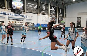 Lanzarote Deportiva captura un partido de balonmano femenino en una pista cubierta, donde jugadoras en uniforme luchan por la pelota en un movimiento de ataque. El estadio cuenta con anuncios publicitarios y espectadores.