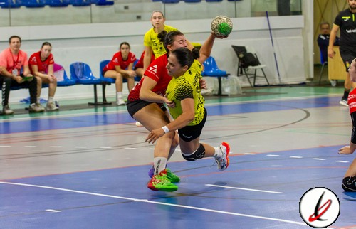 Balón de balonmano en el aire mientras dos jugadoras en acción en una cancha cubierta, con árbitros y equipo en el fondo durante un partido de balonmano en Lanzarote Deportiva.