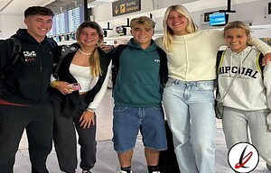 Varios jóvenes en aeropuerto con mochilas y ropa casual, sonrisas y actitud relajada, en el fondo mostradores de facturación y pantallas de información de vuelos, en Lanzarote Deportiva.