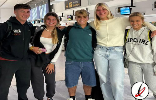 Varios jóvenes en aeropuerto con mochilas y ropa casual, sonrisas y actitud relajada, en el fondo mostradores de facturación y pantallas de información de vuelos, en Lanzarote Deportiva.