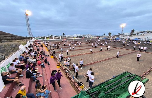 Jugadores y espectadores en un estadio de beach volley en Lanzarote, con actividades en curso y un ambiente animado bajo un cielo nublado.