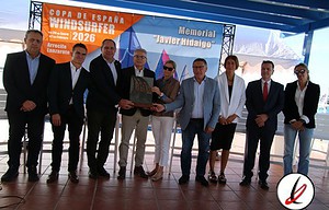 -Un grupo de nueve personas en un evento de homenaje en Lanzarote, con un cartel que indica "Memorial Javier Indiaguo" y "Copa de España Windsurfer 2026".