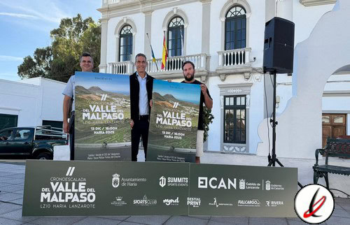 1. Tres hombres en un acto oficial sosteniendo carteles promocionales para una carrera en Lanzarote, en un entorno urbano con edificio histórico y escenario al aire libre.