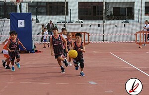 Jóvenes jugadores de baloncesto en una cancha exterior, entrenando y corriendo con un balón de baloncesto, con fondo urbano y red de seguridad en Lanzarote Deporte.