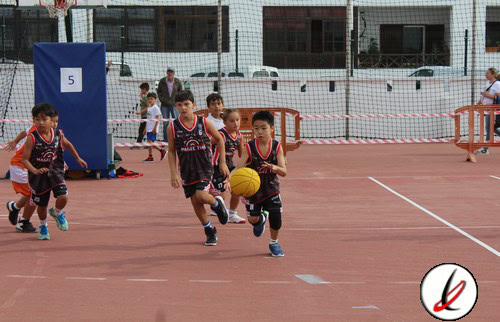 Jóvenes jugadores de baloncesto en una cancha exterior, entrenando y corriendo con un balón de baloncesto, con fondo urbano y red de seguridad en Lanzarote Deporte.